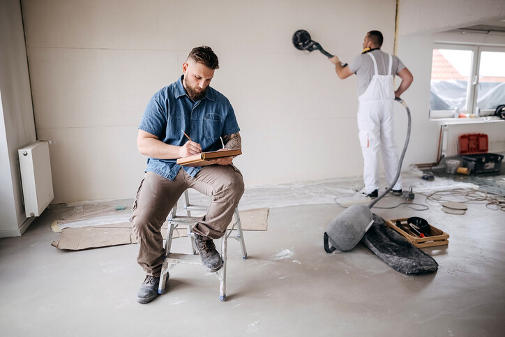 man sting on step ladder writing in book as worker sands drywall in background